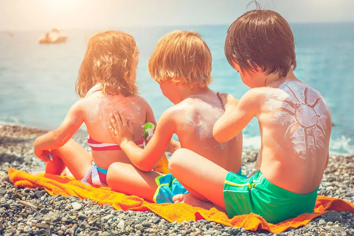 Dzieci na plaży smarują się kremem z filtrem, by uniknąć oparzeń słonecznych. Na plecach jednego z chłopców namalowano słońce.