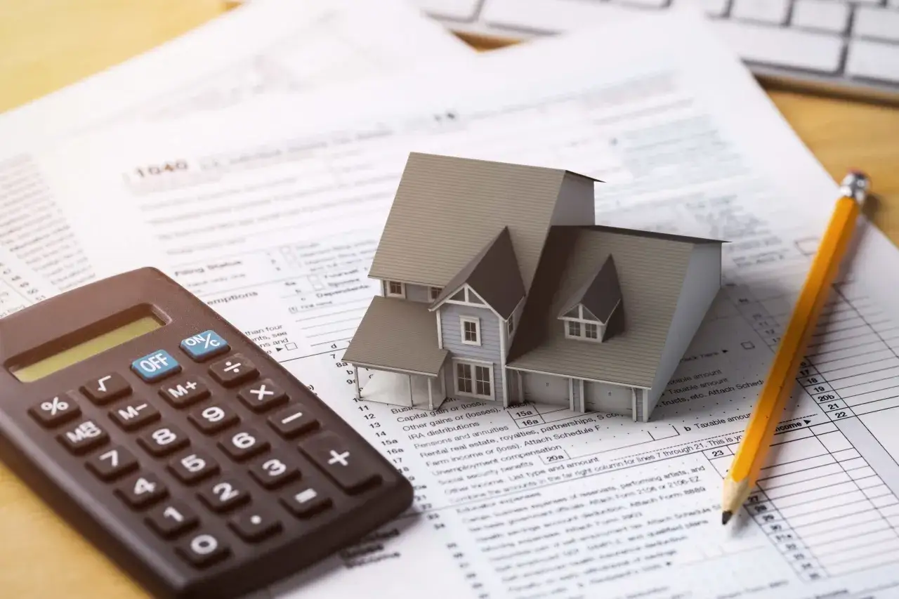 Casa modelo sobre formulario 1040, calculadora y l&aacute;piz. El heredero no paga gastos vivienda, pero debe declarar ingresos.
