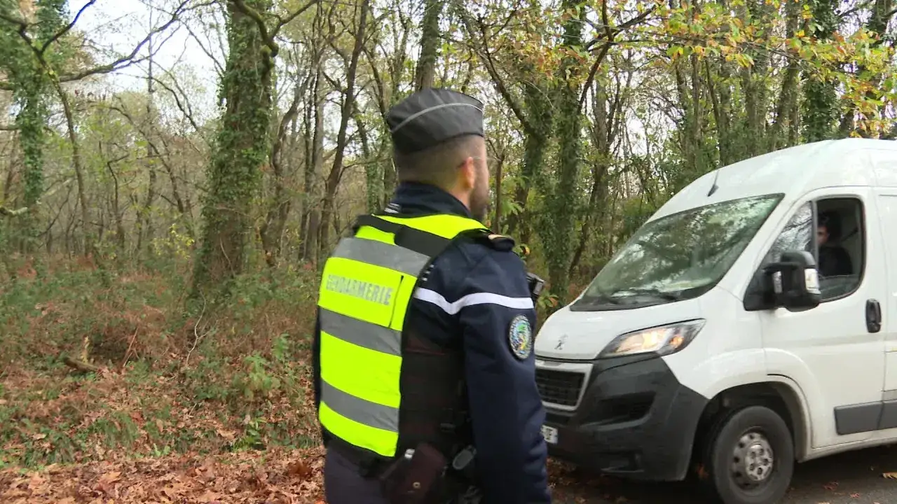 Un gendarme en uniforme fluorescent observe un véhicule blanc. Le conducteur est au volant, peut-être distrait par son téléphone.