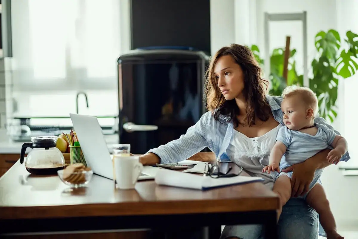 Młoda mama z dzieckiem na rękach pracuje przy laptopie. Czy na macierzyńskim można pracować? Tak, jak widać na zdjęciu.