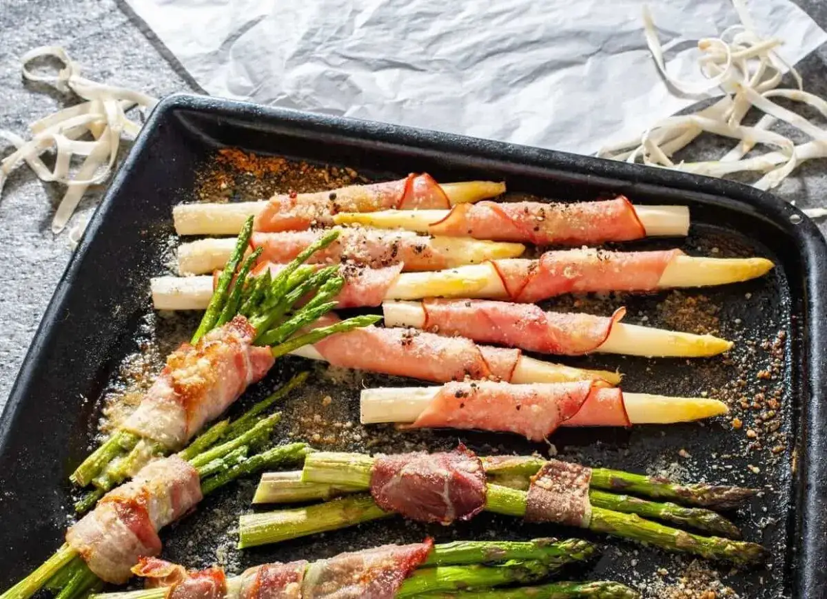 Zielone i białe szparagi owinięte szynką parmeńską, posypane przyprawami, gotowe do pieczenia.