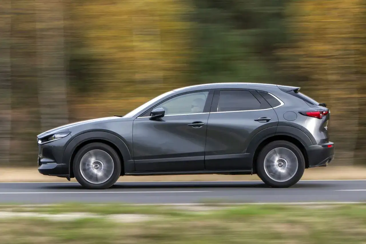 Szary Mazda CX-30 w ruchu, z rozmytym tłem drzew. Kierowca w okularach prowadzi auto po drodze.