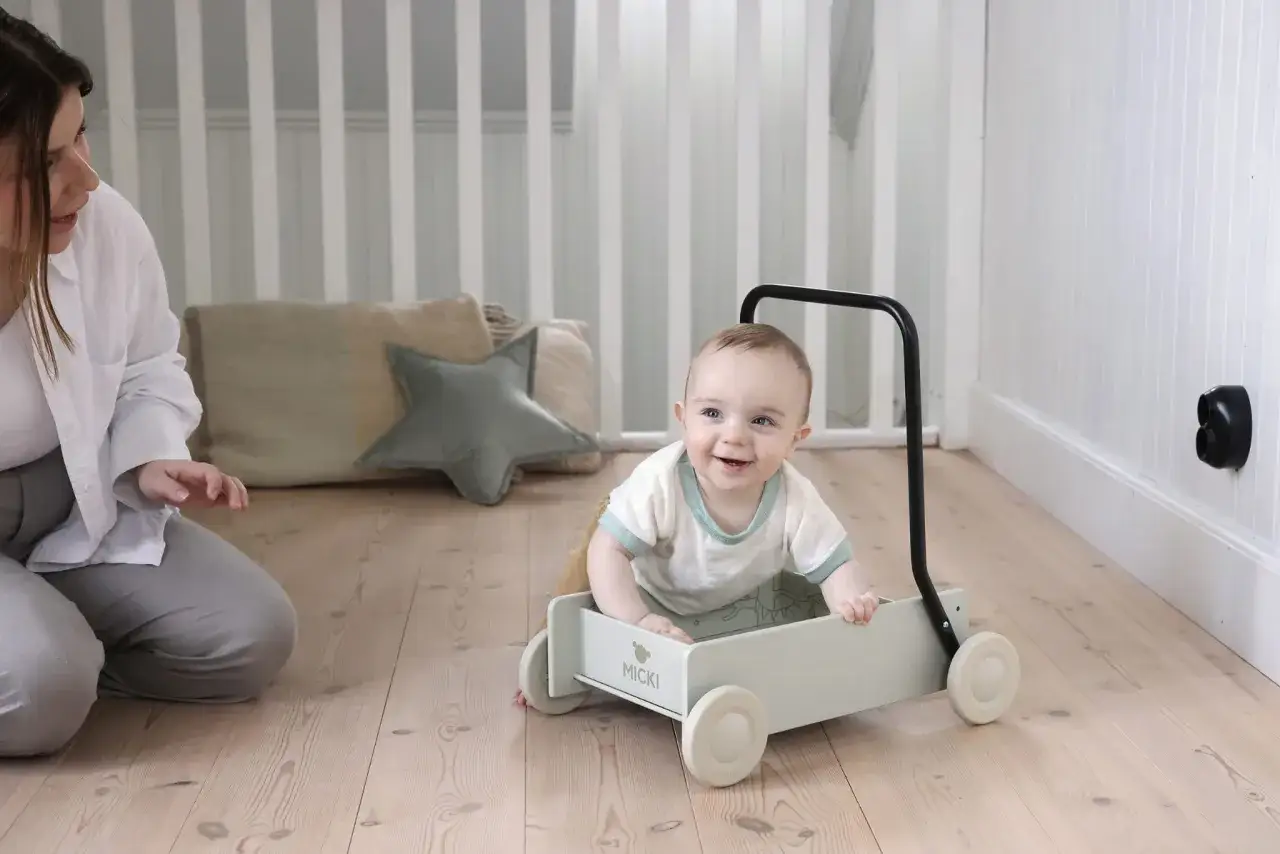 Uśmiechnięte dziecko w pchaczu wspiera fizjoterapię i pierwsze kroki. Mama czuwa obok.