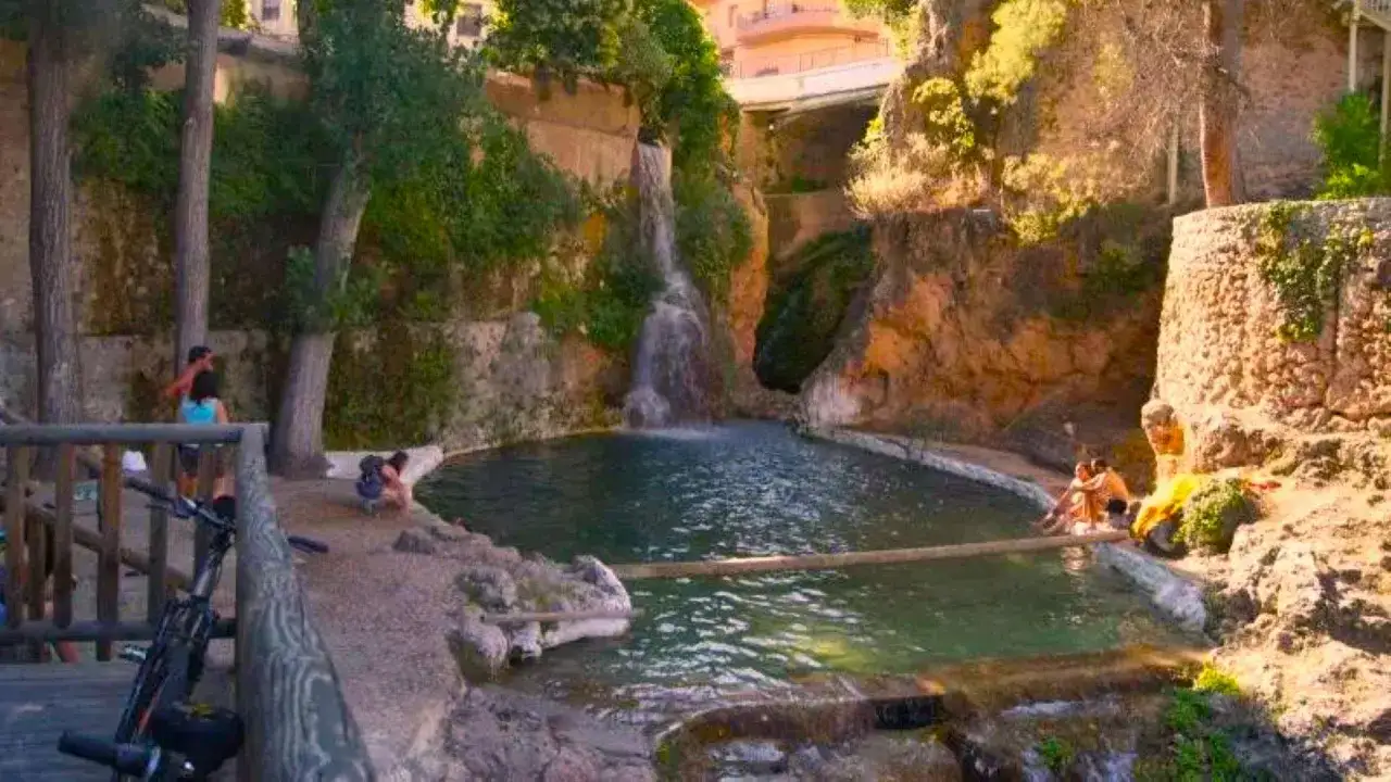 Una cascada alimenta la letur piscina natural, donde unas personas disfrutan del agua rodeadas de rocas y vegetación.