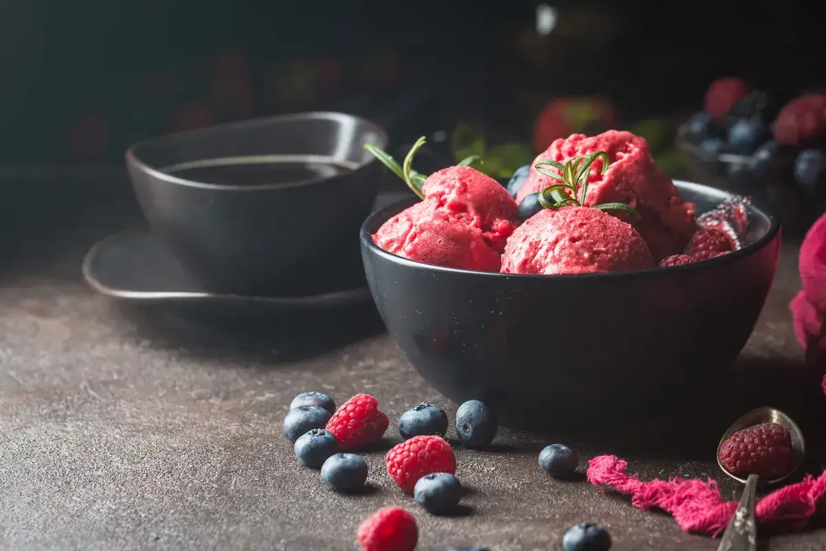 Aksamitny sorbet malinowy z jagodami i rozmarynem, obok filiżanka kawy. Idealny deser na upalne dni.