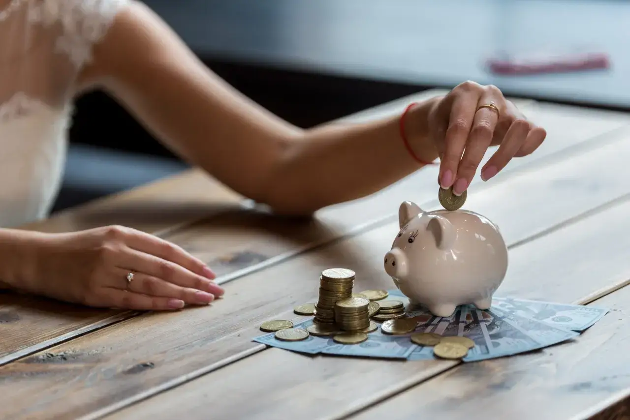 Kobieta wkłada monetę do świnki skarbonki. Obok leżą stosy monet i banknoty. Oszczędzanie na przyszłość, może na opłatę ZAIKS.