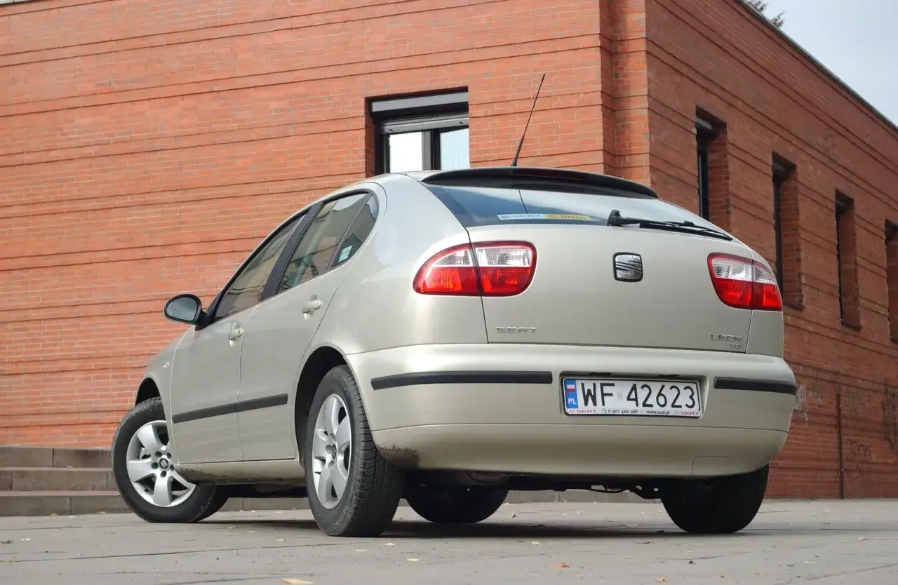 Złoty Seat Leon 1 z widocznymi żarówkami tylnych świateł, zaparkowany przy ceglanym budynku.