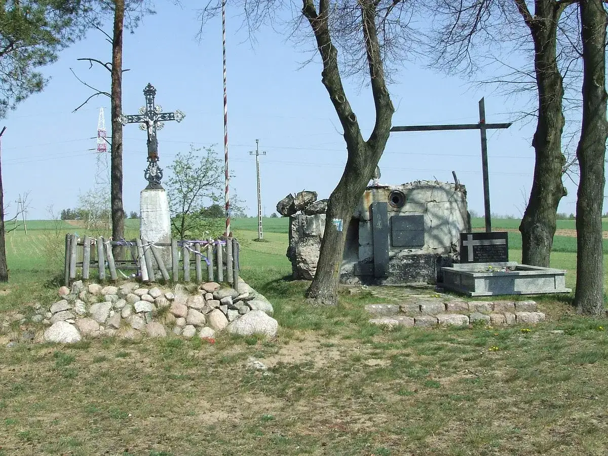 Kamienny krzyż i pomnik z tablicami, otoczone kamieniami i drewnianym płotkiem. W tle drzewa i pola. Wstążki w kolorach tęczy, jak jedwabne, zdobią płotek.
