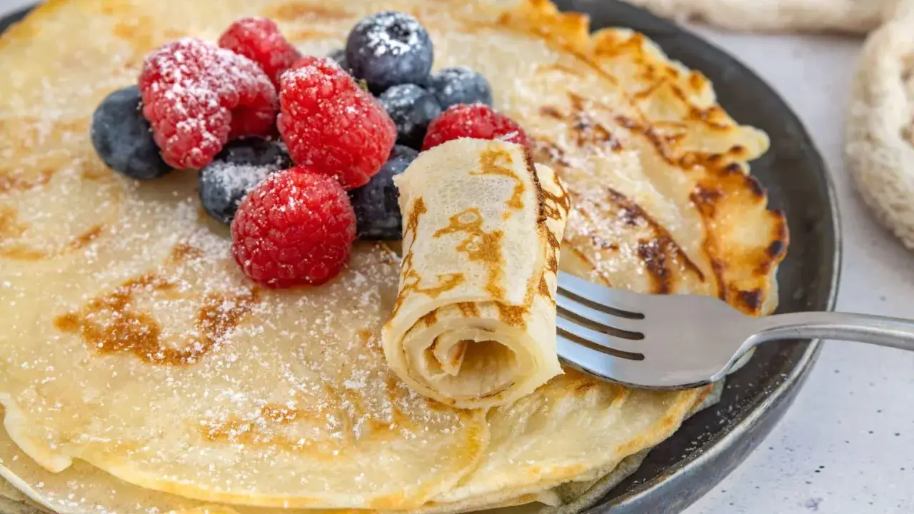 Une assiette de cr&ecirc;pes moelleuses, garnies de framboises et myrtilles, saupoudr&eacute;es de sucre glace. Une cr&ecirc;pe est roul&eacute;e sur une fourchette.