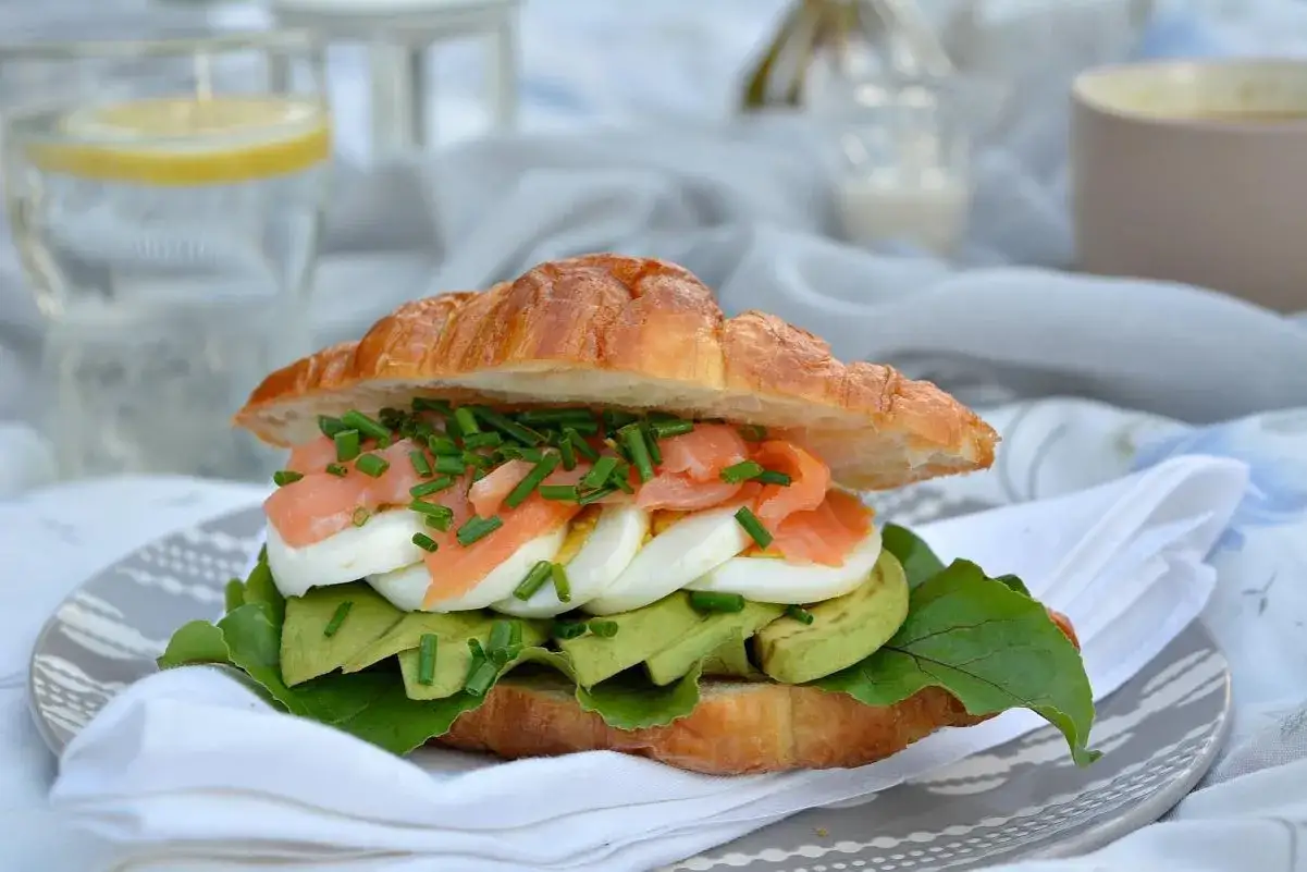 Pyszna kanapka z łososiem, awokado i jajkiem na miękkim rogaliku, posypana szczypiorkiem. Idealna na lekki lunch.