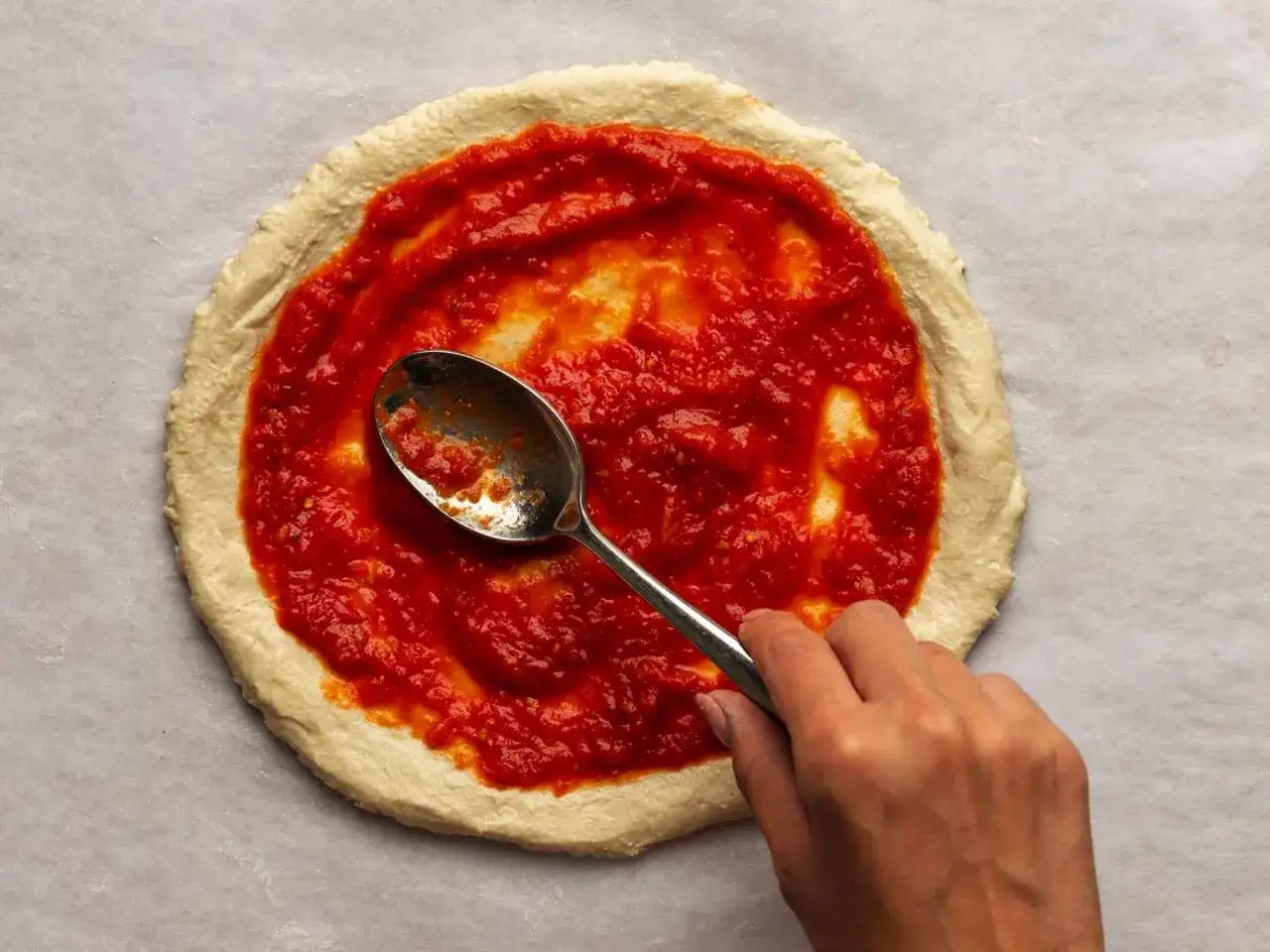 Ręka rozsmarowuje łyżką pyszny, gęsty sos do pizzy na surowym cieście.