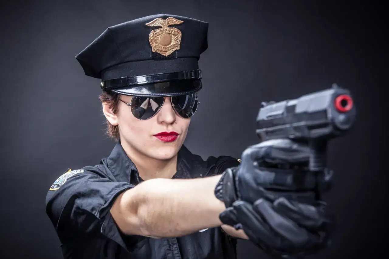Policjantka w czapce i okularach przeciwsłonecznych celuje z pistoletu. Jej dłonie w rękawiczkach pewnie trzymają broń, gotową do użycia a wykorzystania w każdej chwili.
