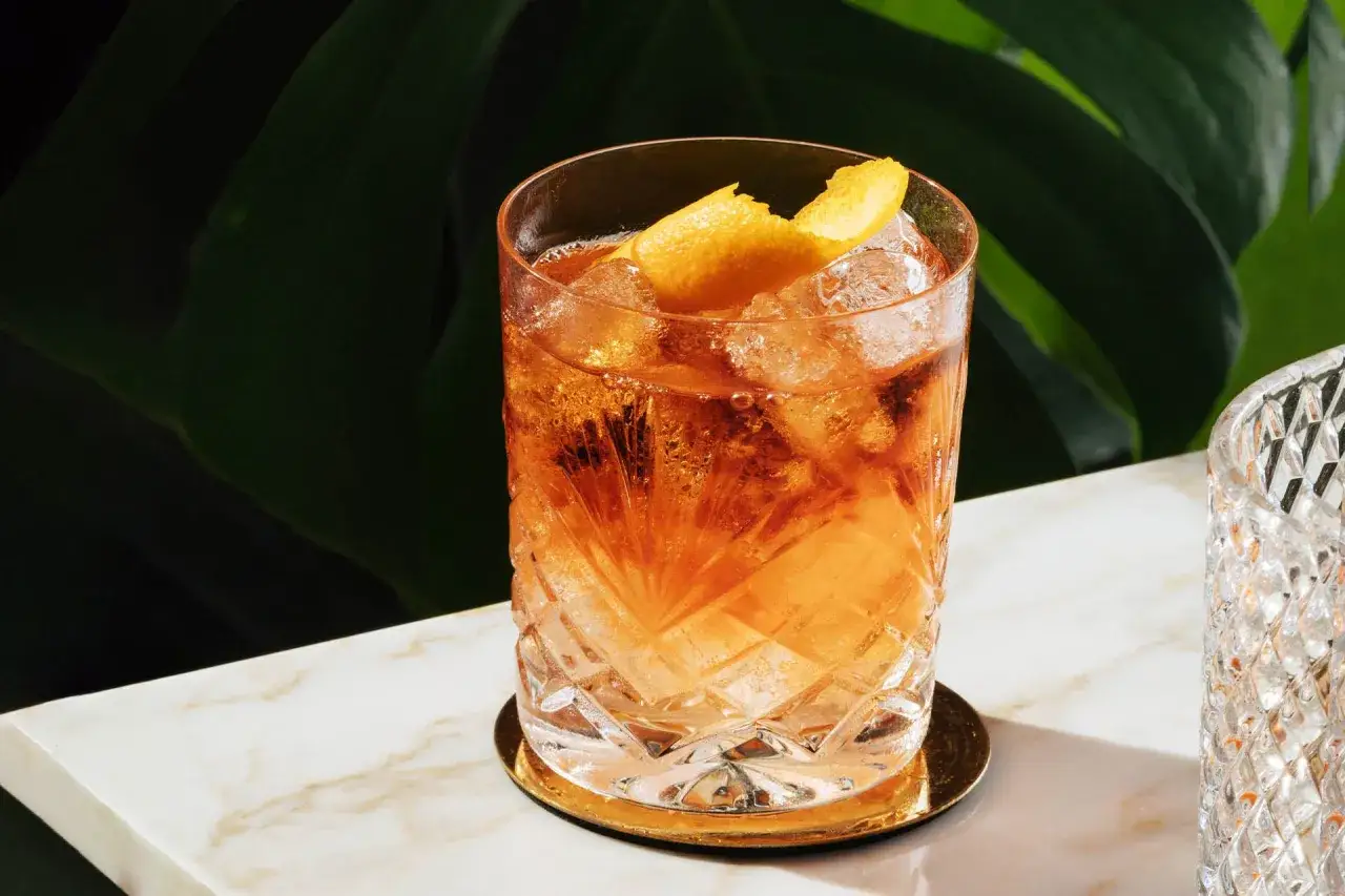 Orzeźwiający Negroni Sbagliato z pomarańczą i lodem, podany w szklance z fasetowanym szkłem, na marmurowym blacie.