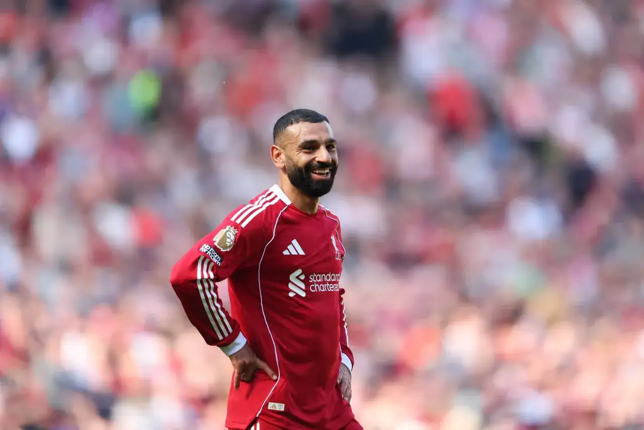 Uśmiechnięty piłkarz w czerwonej koszulce Liverpoolu, gdzie gra Salah, z ręką na biodrze, na tle rozmytych kibiców.