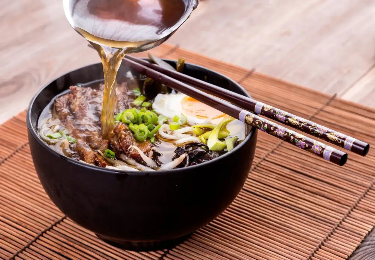 Un tazón humeante de comida asiática, ramen con carne, huevo y cebolleta, se sirve con palillos decorados.