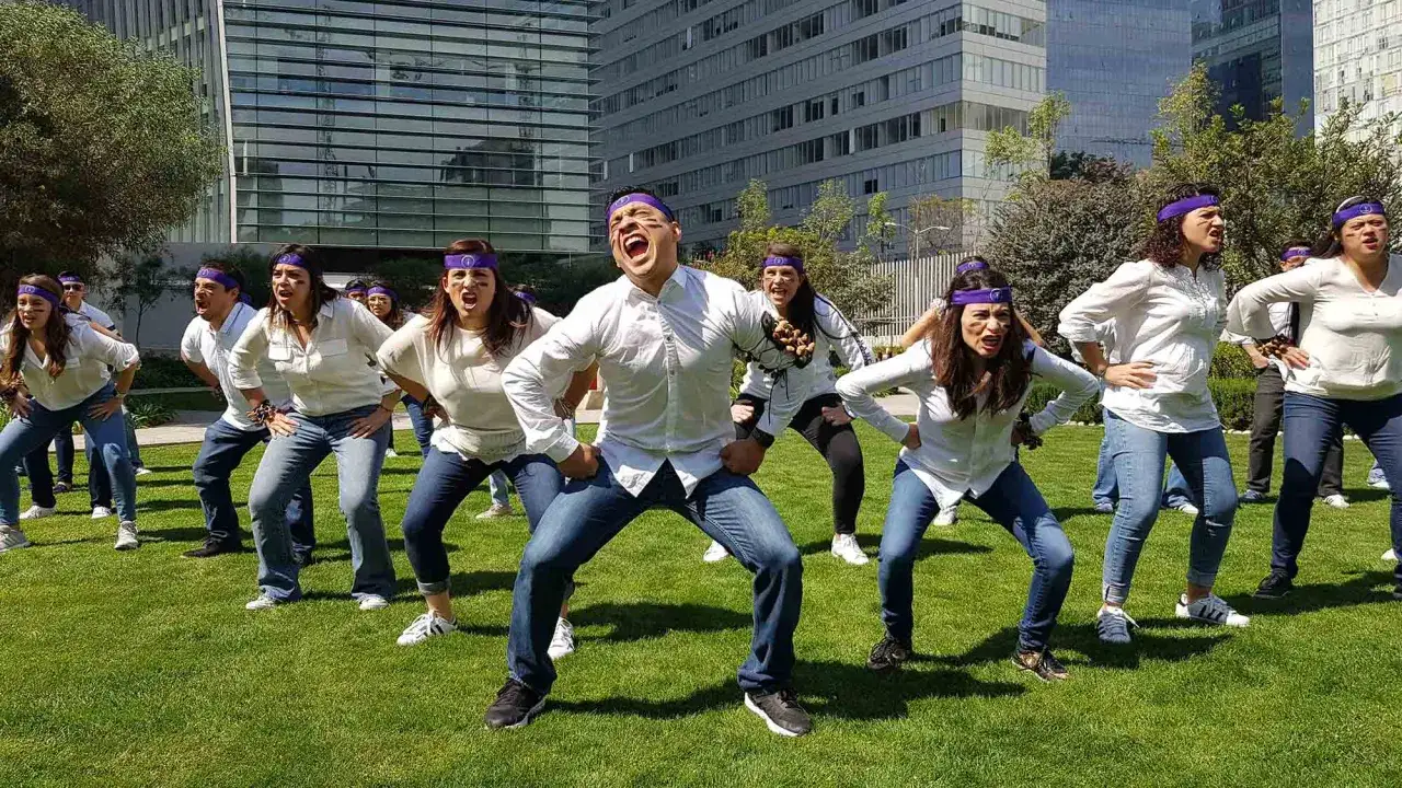 Grupo de personas en actividades para empresas team building, con vinchas púrpuras y ropa blanca, bailando en un césped frente a edificios modernos.