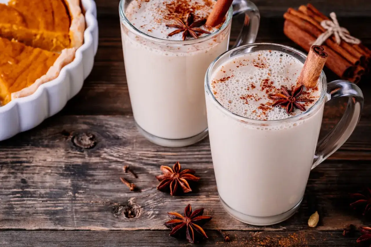 Dwa kubki aromatycznego chai latte, ozdobione cynamonem i anyżem, obok kawałka dyniowego placka. Idealne na jesienne popołudnie.