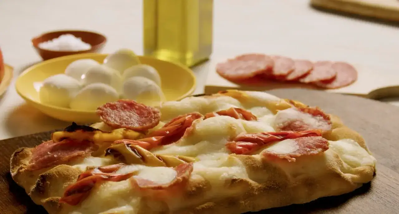 En härlig pinsa med salami, mozzarella och rostade tomater. Bredvid syns små mozzarellabollar, salt och olivolja.