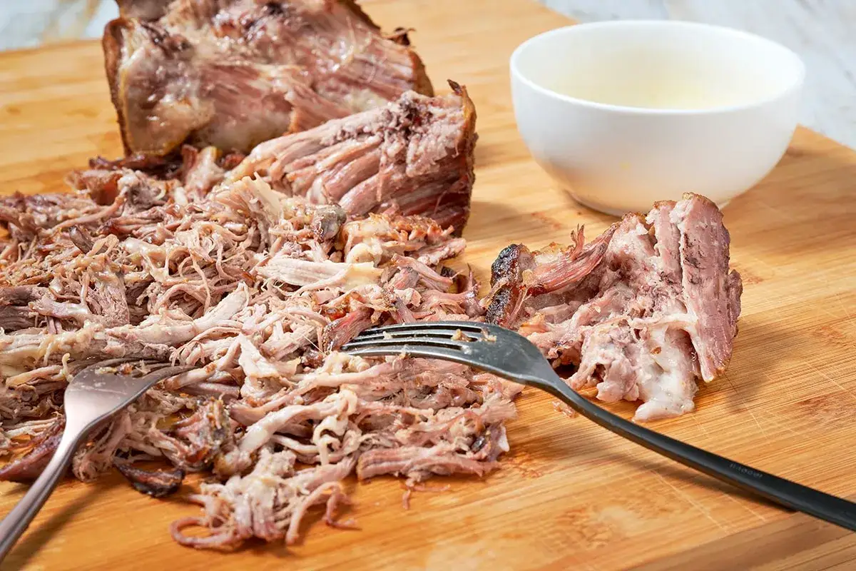Carne di maiale sfilacciata, pronta per la tua pulled pork ricetta. Due forchette la lavorano su un tagliere di legno, accanto a una ciotolina.