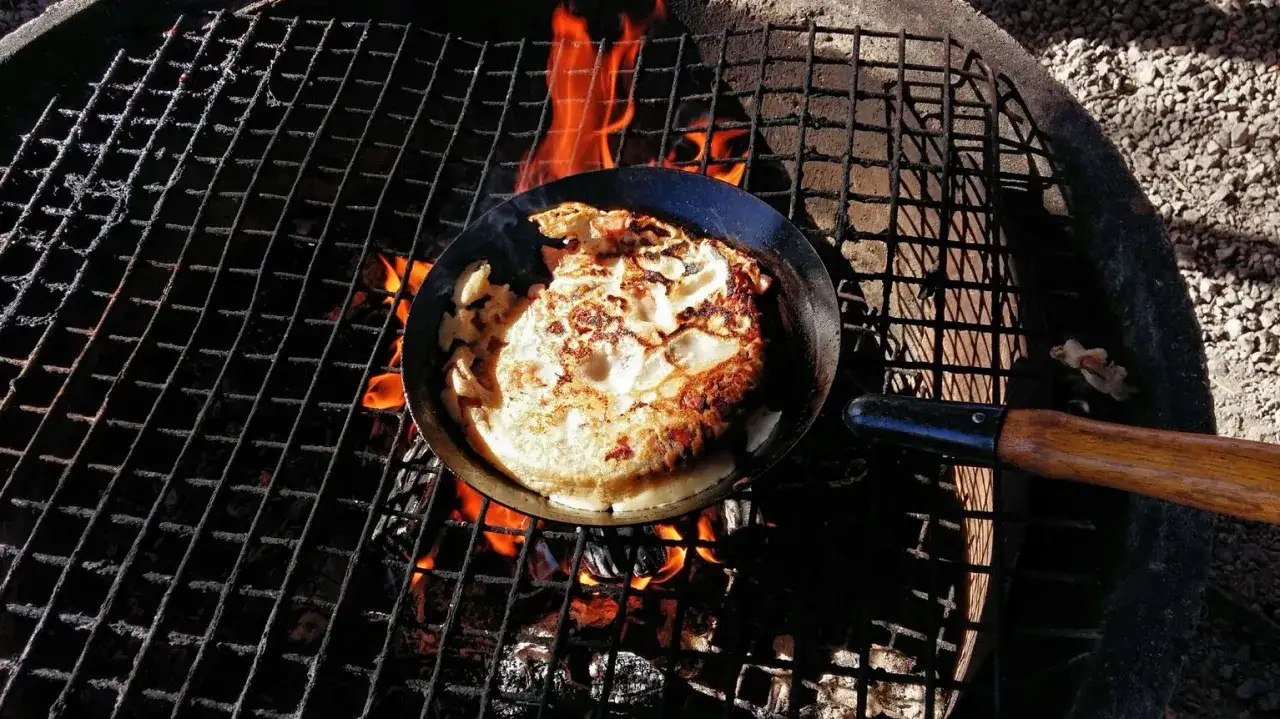 Kolbulle smet steks i stekpanna över öppen eld på en grill. Flammor syns under gallret.