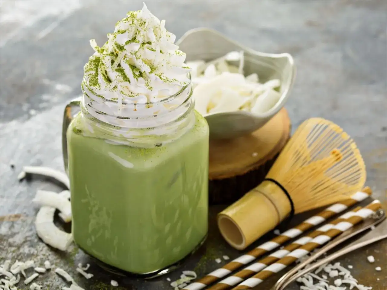 Orzeźwiające matcha z mlekiem kokosowym, udekorowane bitą śmietaną i wiórkami kokosowymi. Obok bambusowa miotełka do matchy i słomki.