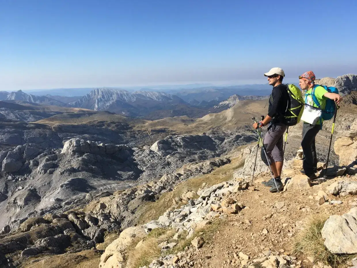 Dos excursionistas disfrutan de la vista panorámica de la transpirenaica, con picos montañosos y un cielo azul despejado.