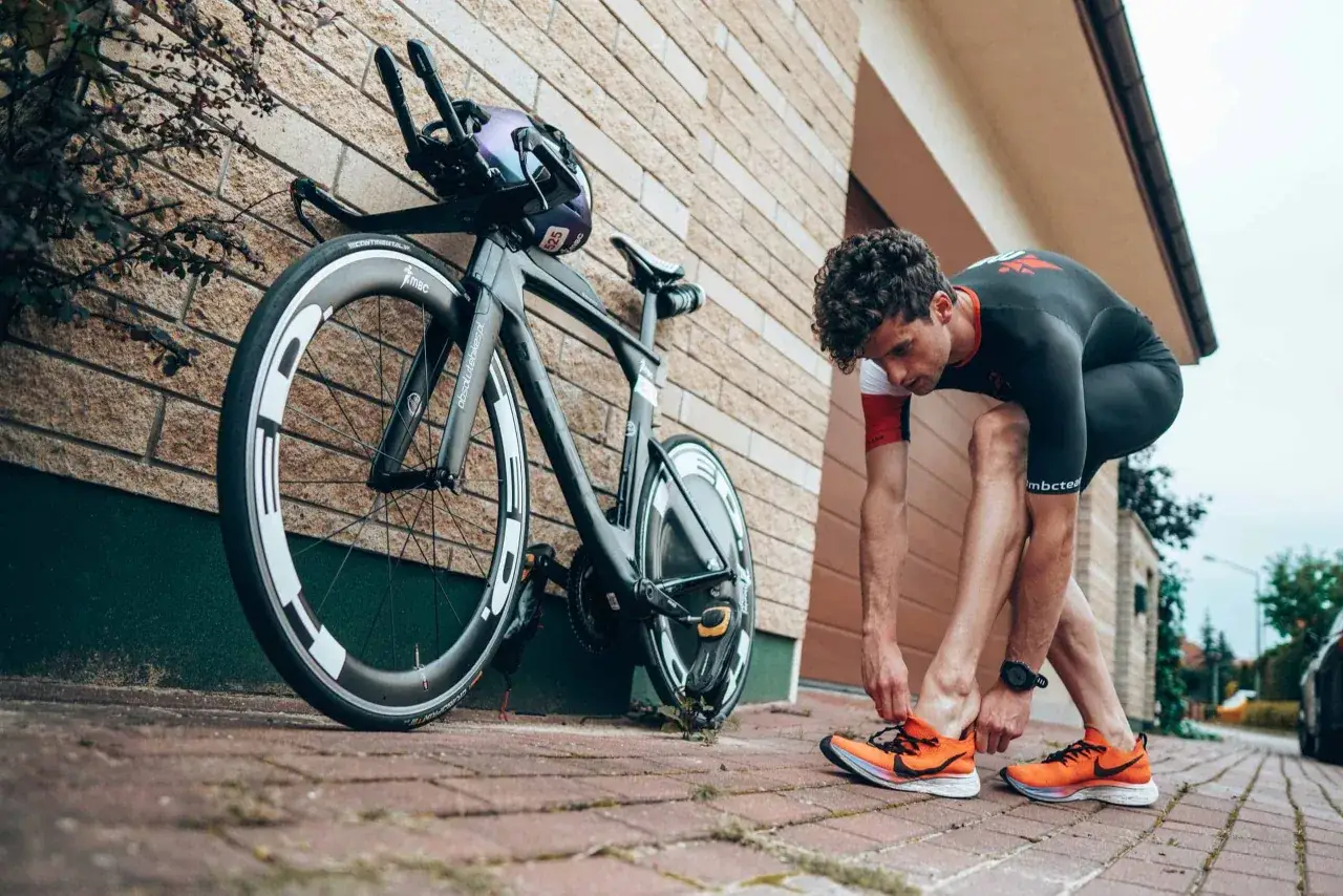 Zawodnik w stroju do triathlonu olimpijskiego sznuruje pomarańczowe buty obok roweru.