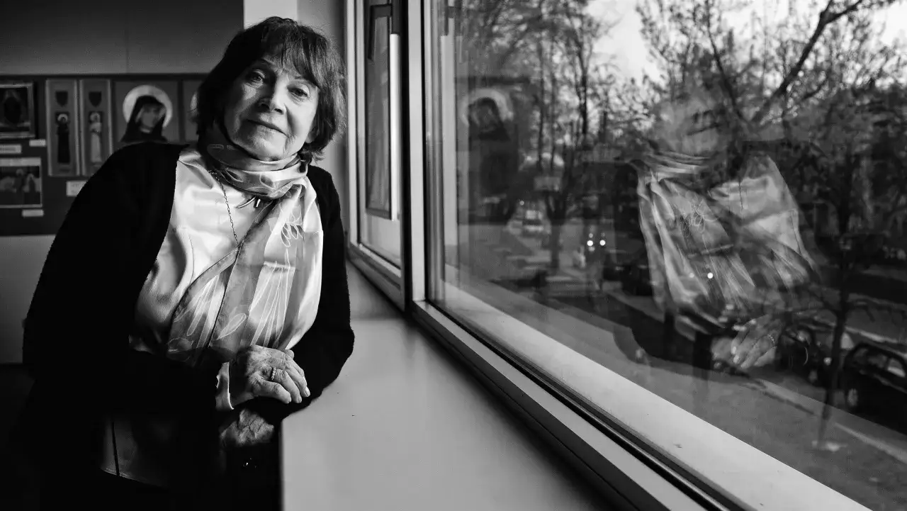 Alicja Pawlicka, zamyślona, przy oknie. W tle obrazy, a za szybą odbija się świat.
