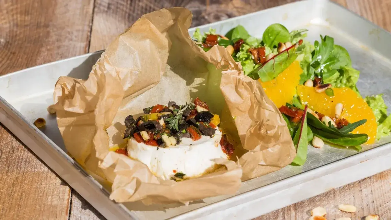 Leckeres vegan grillen: Ein runder, weißer Block, umhüllt von Backpapier, belegt mit Oliven und Tomaten, daneben ein frischer Salat mit Orangenscheiben.