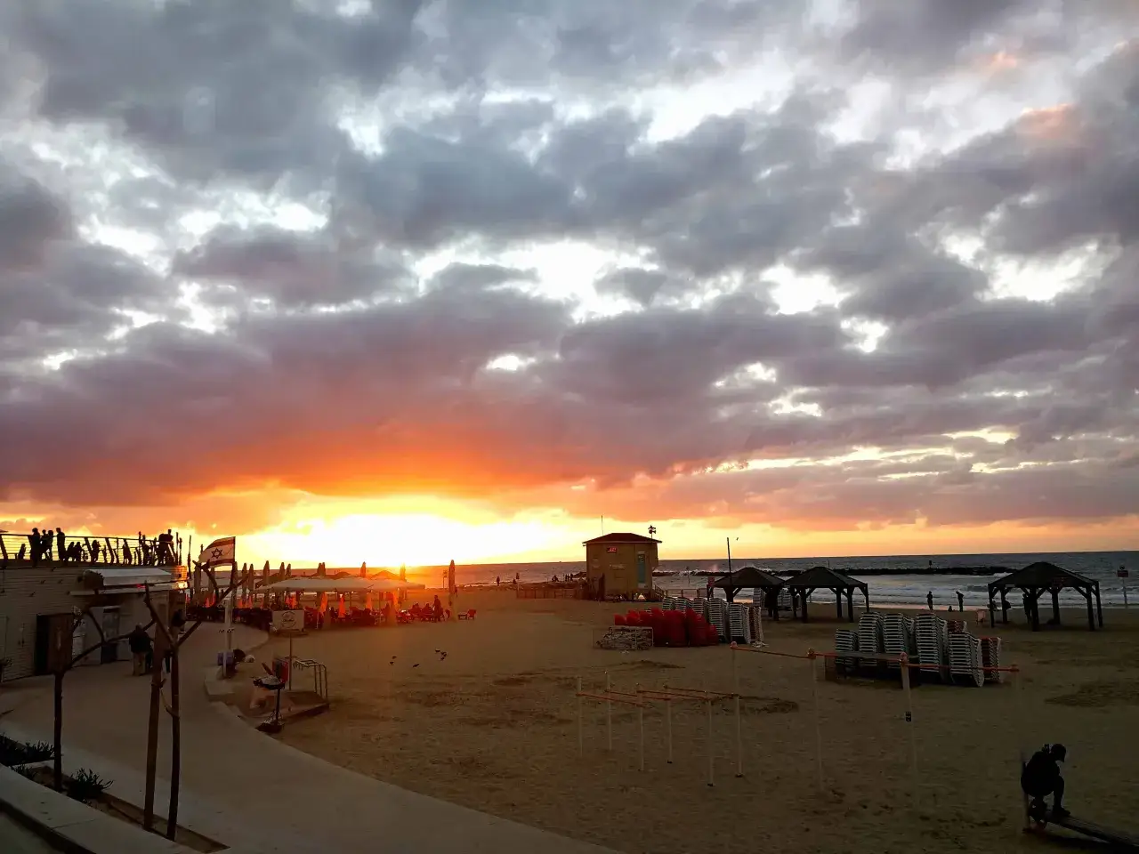 Spektakul&auml;rer Sonnenuntergang &uuml;ber dem Strand von Tel Aviv. Wolken f&auml;rben sich rot und orange, w&auml;hrend Menschen auf der Promenade stehen und den Ausblick genie&szlig;en.