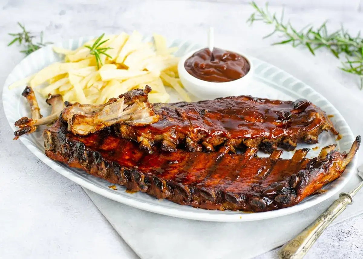 Costillas de cerdo al horno cubiertas de salsa barbacoa, servidas con patatas fritas y una ramita de romero.