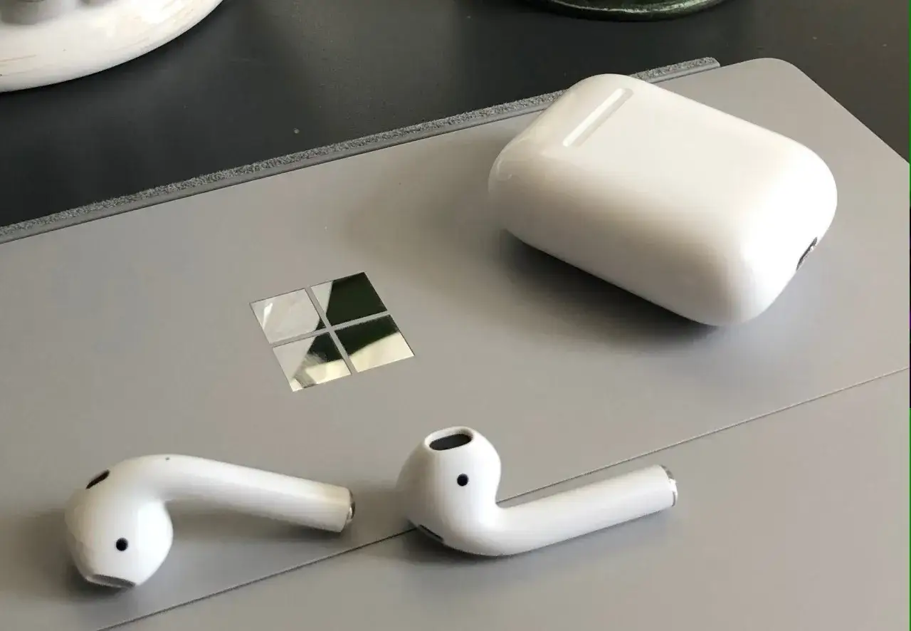 AirPods i etui na szarym laptopie z logo Windows. Dowiedz się, jak podłączyć AirPods do Windowsa.