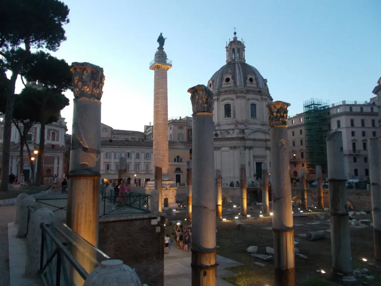 Antiche colonne romane illuminate al crepuscolo, con una maestosa colonna traiana e una chiesa con cupola sullo sfondo.