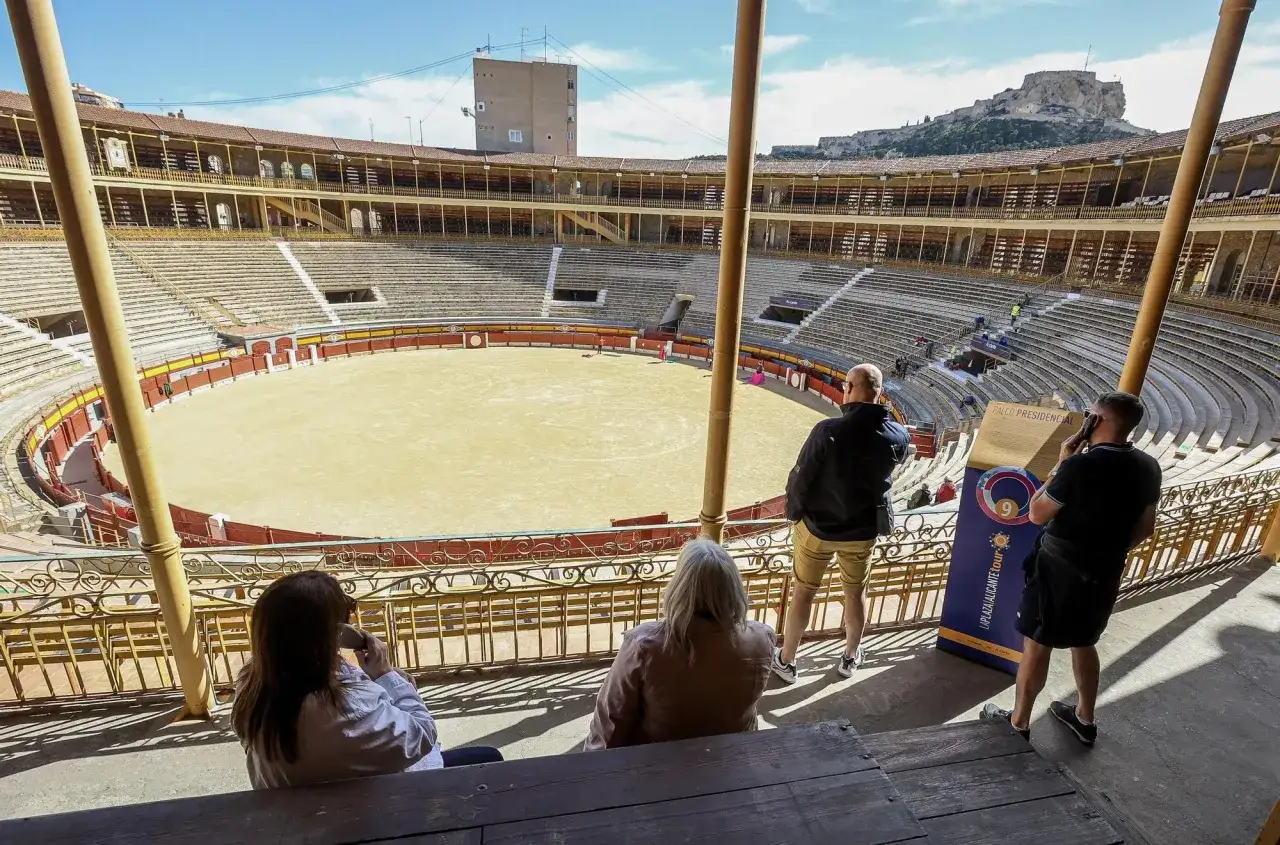 Vista de la plaza de toros de Alicante, con el aforo vac&iacute;o y turistas observando. Al fondo, el castillo de Santa B&aacute;rbara.