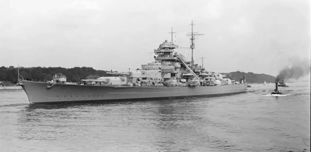 Potężny pancernik Bismarck na wodzie, z widocznymi działami i nadbudówkami. W tle widać zielone wybrzeże i małe statki.