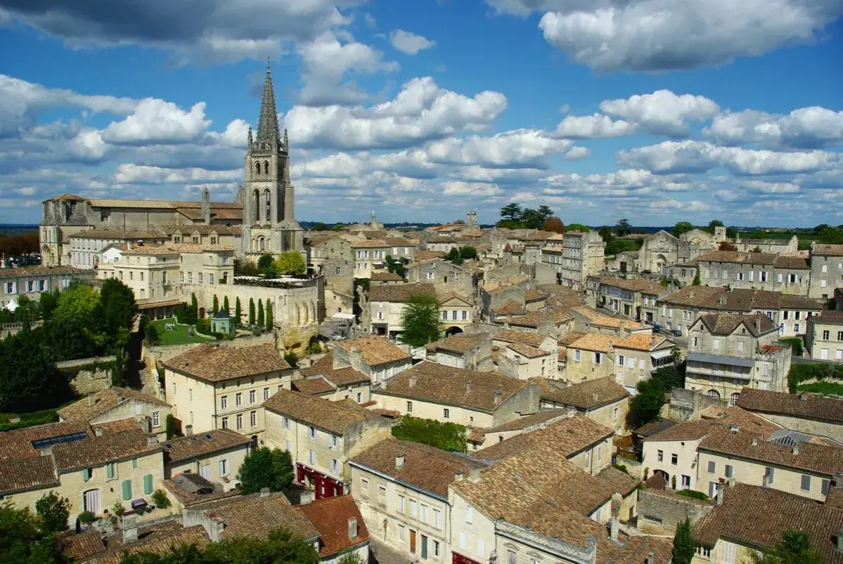 Vue panoramique de Saint-Émilion, avec son église imposante et ses toits de tuiles ocres sous un ciel bleu parsemé de nuages blancs. Une belle journée pour une visite.