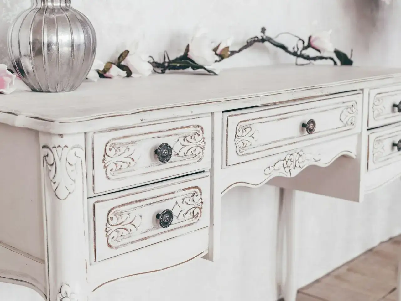 Biała, postarzana komoda w stylu shabby chic z ozdobnymi uchwytami i rzeźbieniami. Na blacie wazon i gałązka z kwiatami.