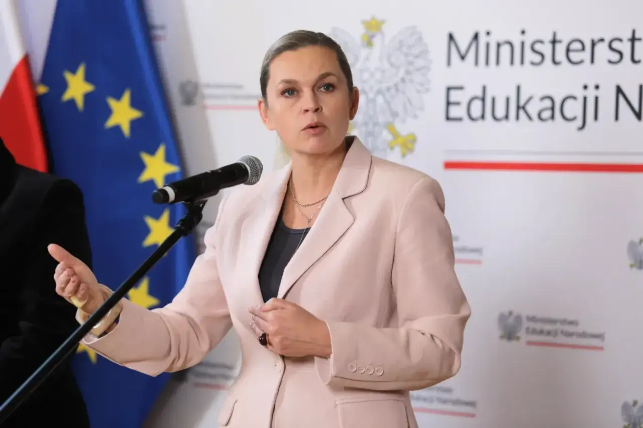 Kobieta w różowym żakiecie przemawia przy mikrofonie, na tle flagi Polski i UE. Może być oskarżona w procesie sądowym.
