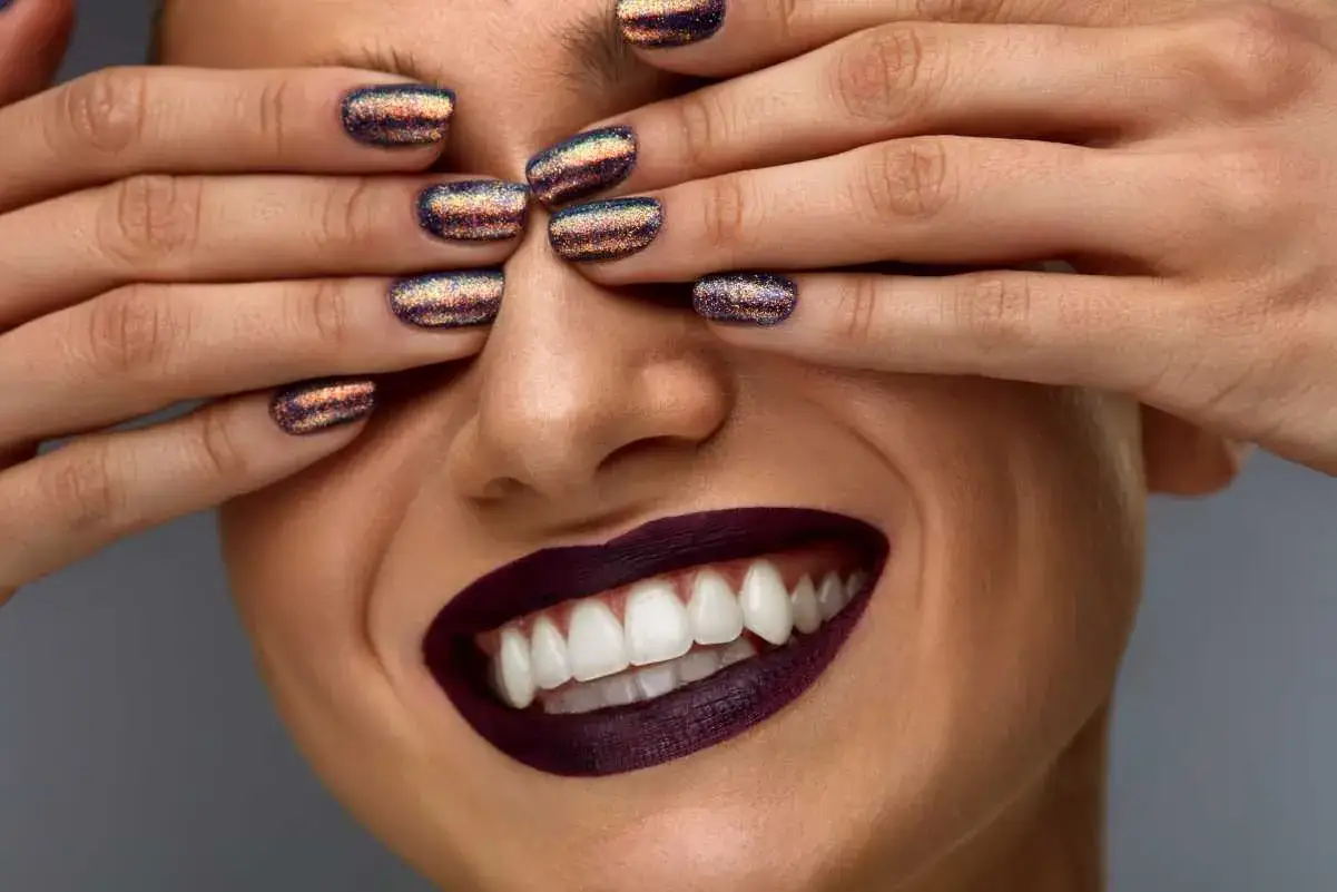 Unghie per l'inverno scintillanti, con un tocco di viola e oro, coprono gli occhi di una donna che sorride mostrando labbra scure e denti bianchi.