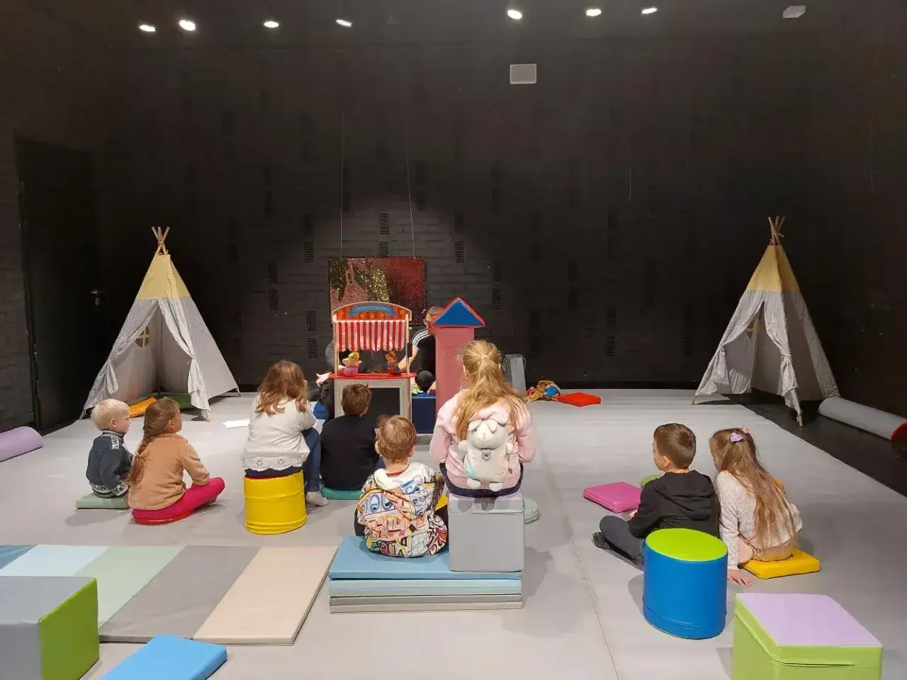 Dzieci siedzą na podłodze, oglądając przedstawienie w teatrze dla młodzieży. W tle namioty tipi i scena kukiełkowa.