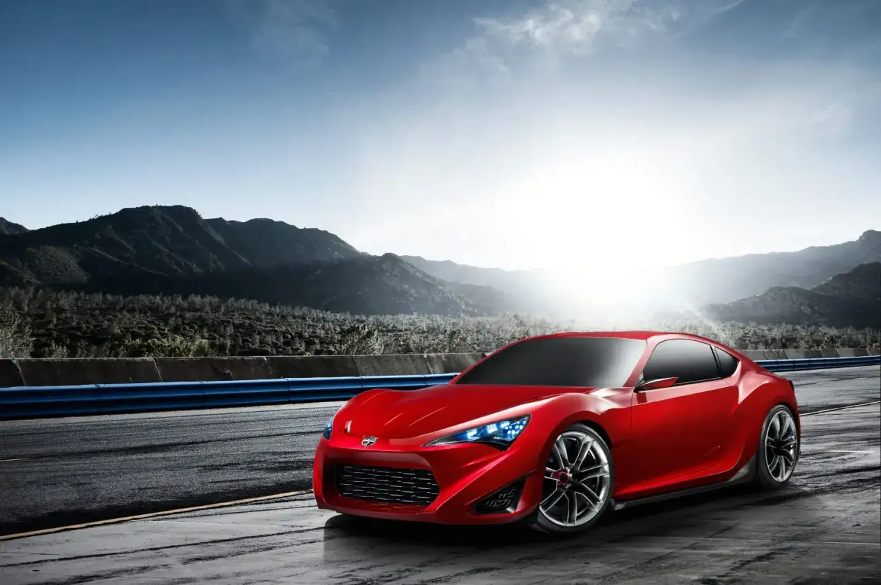 Czerwony Scion FR-S na torze wyścigowym, z górami w tle i słońcem przebijającym się przez chmury.