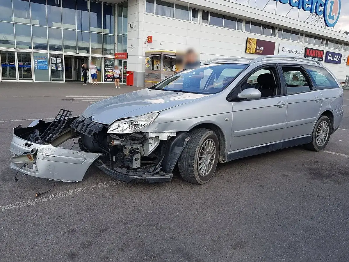 Srebrne kombi z uszkodzonym przodem po kolizji na terenie prywatnym przed supermarketem.
