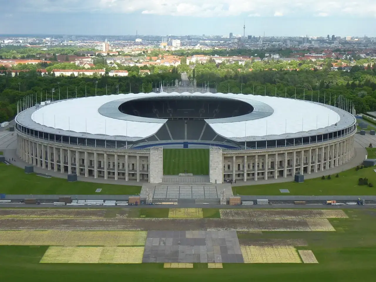 Das Olympiastadion Berlin mit seinem markanten Dach und der Skyline im Hintergrund. In der Ferne ragt der Fernsehturm, der wie ein glockenturm Berlin &uuml;berragt.