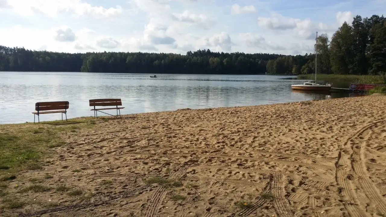 Piękny dzień nad jeziorem. Dwa ławki na piaszczystej plaży zapraszają do odpoczynku. W oddali żaglówka i kajak. **Tleń atrakcje** czekają!