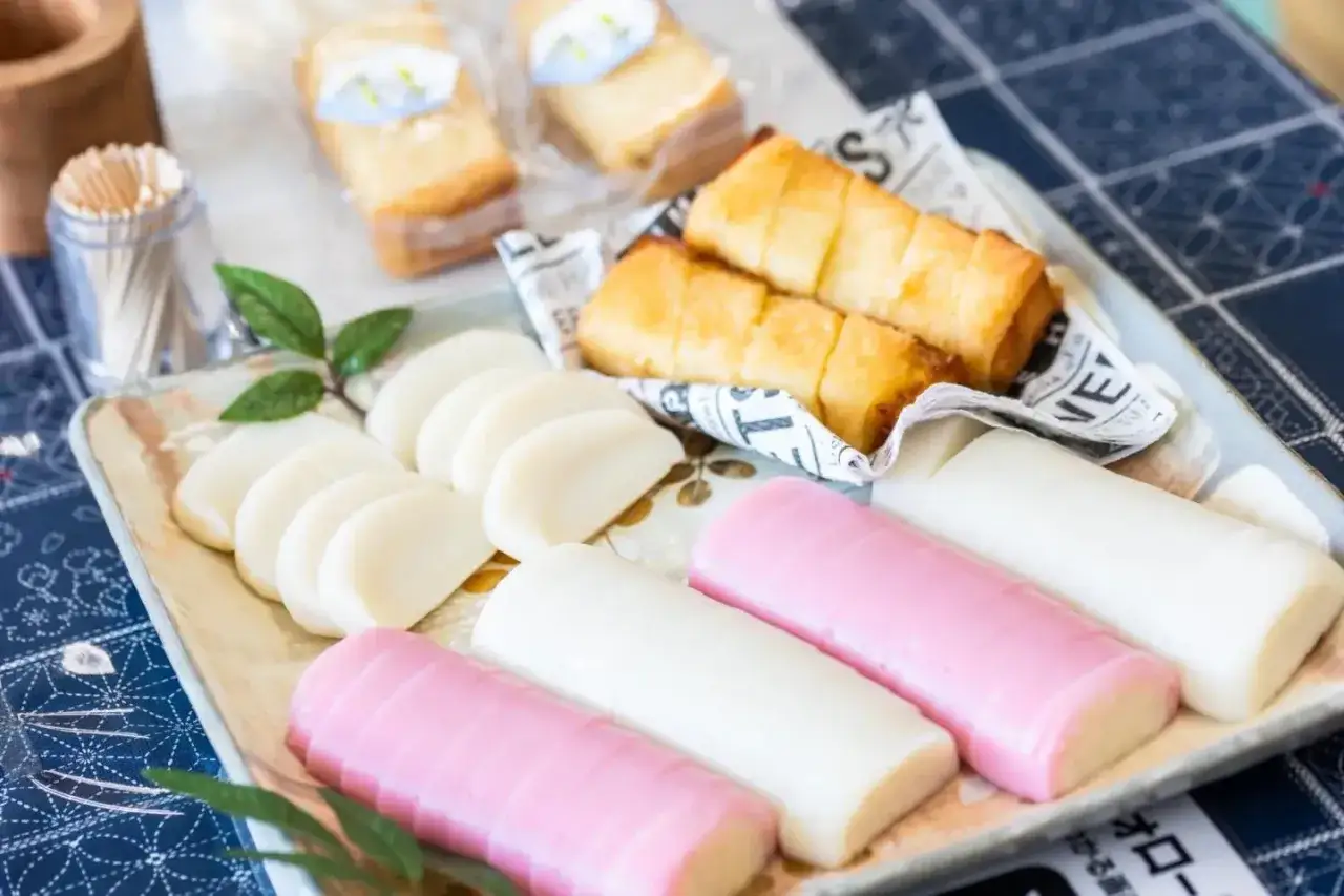 A platter of colorful Japanese fish cake, including sliced white pieces and pink and white logs, with fried rolls in the background.