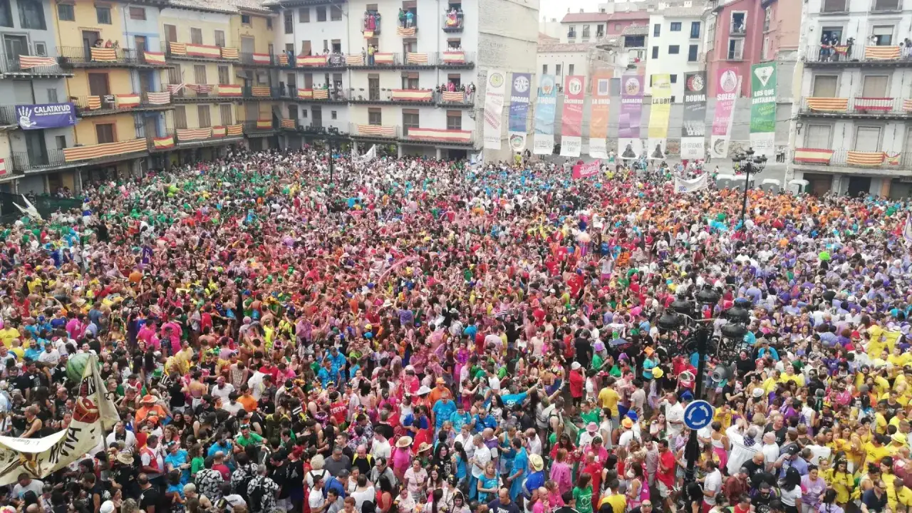Multitud de gente vestida de colores vibrantes celebra las fiestas de Calatayud en una plaza rodeada de edificios decorados con banderas.