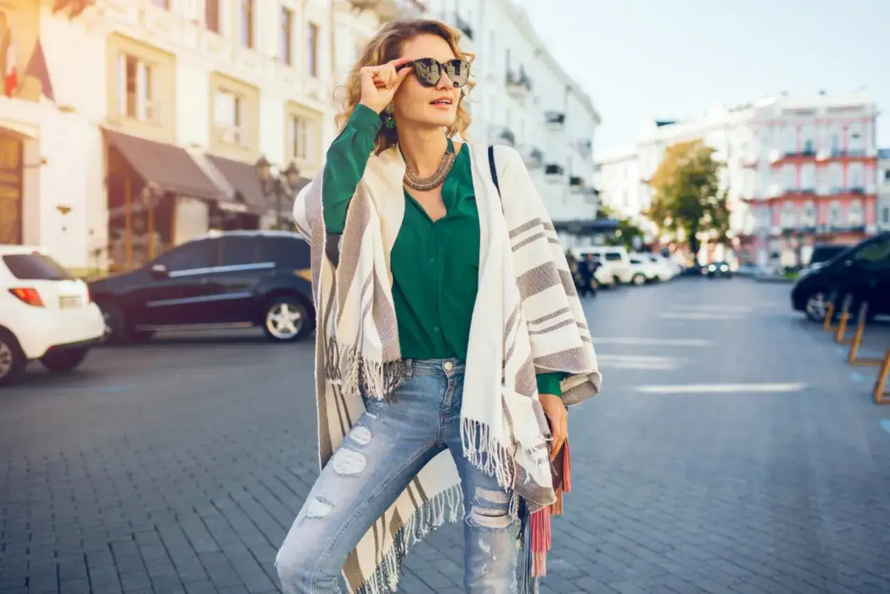 Mujer de 40 a&ntilde;os con look casual, gafas de sol, blusa verde, jeans rotos y un poncho a rayas.