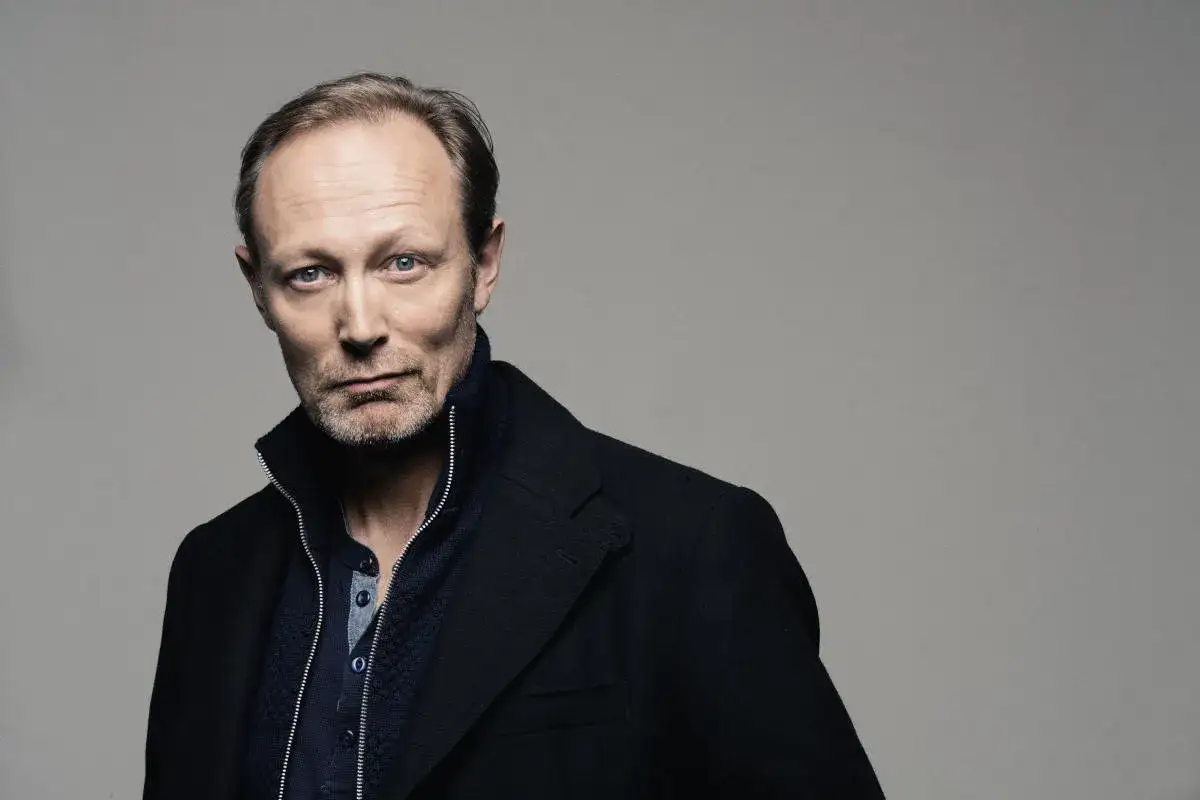Porträtt av skådespelaren Lars Mikkelsen, känd från filmer och tv-program. Han bär en mörk rock över en blå tröja.