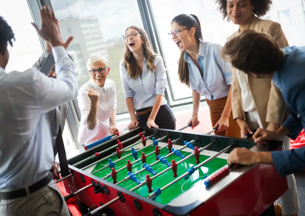 A diverse group of colleagues celebrates a win during employee engagement games of foosball, their laughter echoing in the bright office.