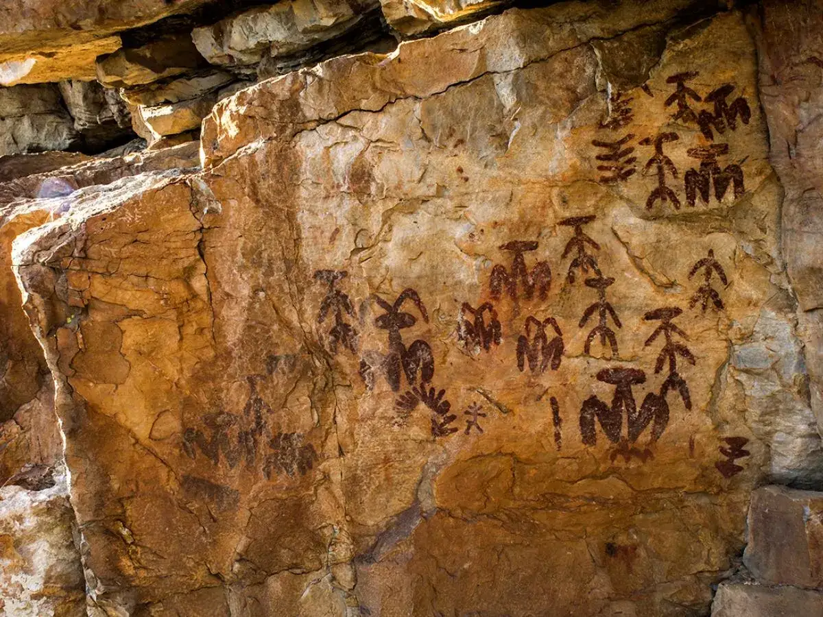 Antiguos grabados en roca muestran diversos tipos de pinturas rupestres, con figuras antropomorfas y zoomorfas en tonos rojizos sobre la pared de una cueva.
